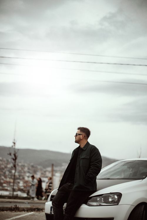 Free Man in black outfit leaning on a car, gazing away on a cloudy day. Stock Photo