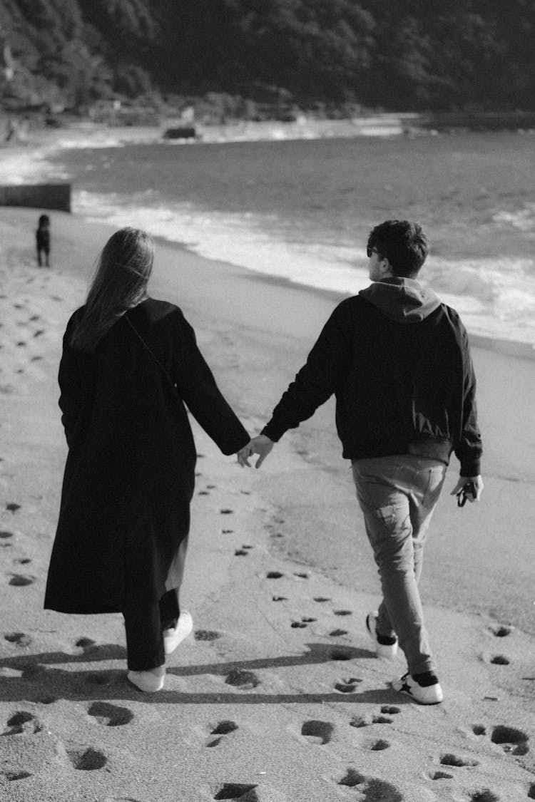 Black And White Photo Of A Young Couple Walking On The Beach Holding Hands