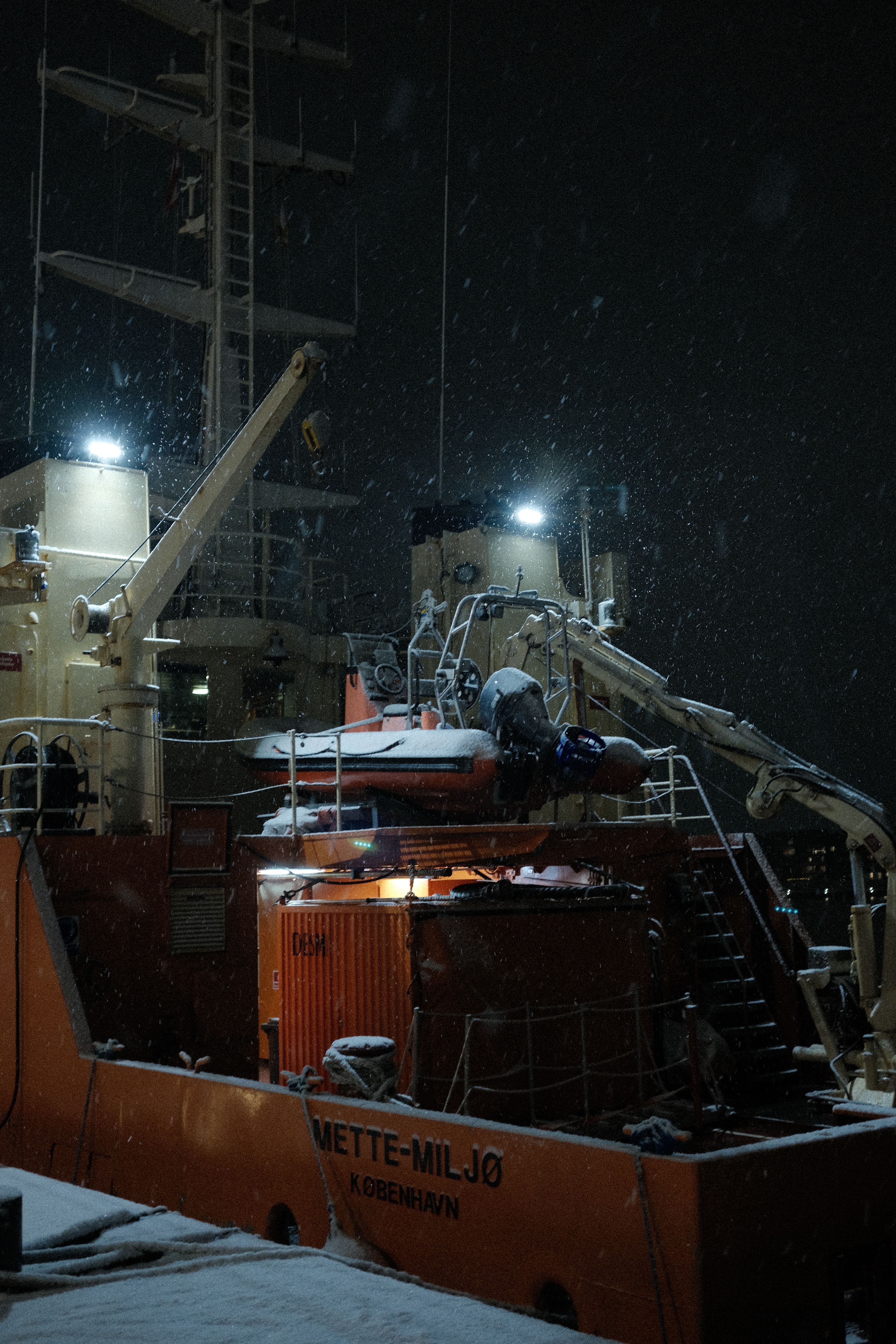Illuminated ship deck covered in snow, at night, with heavy equipment on board.