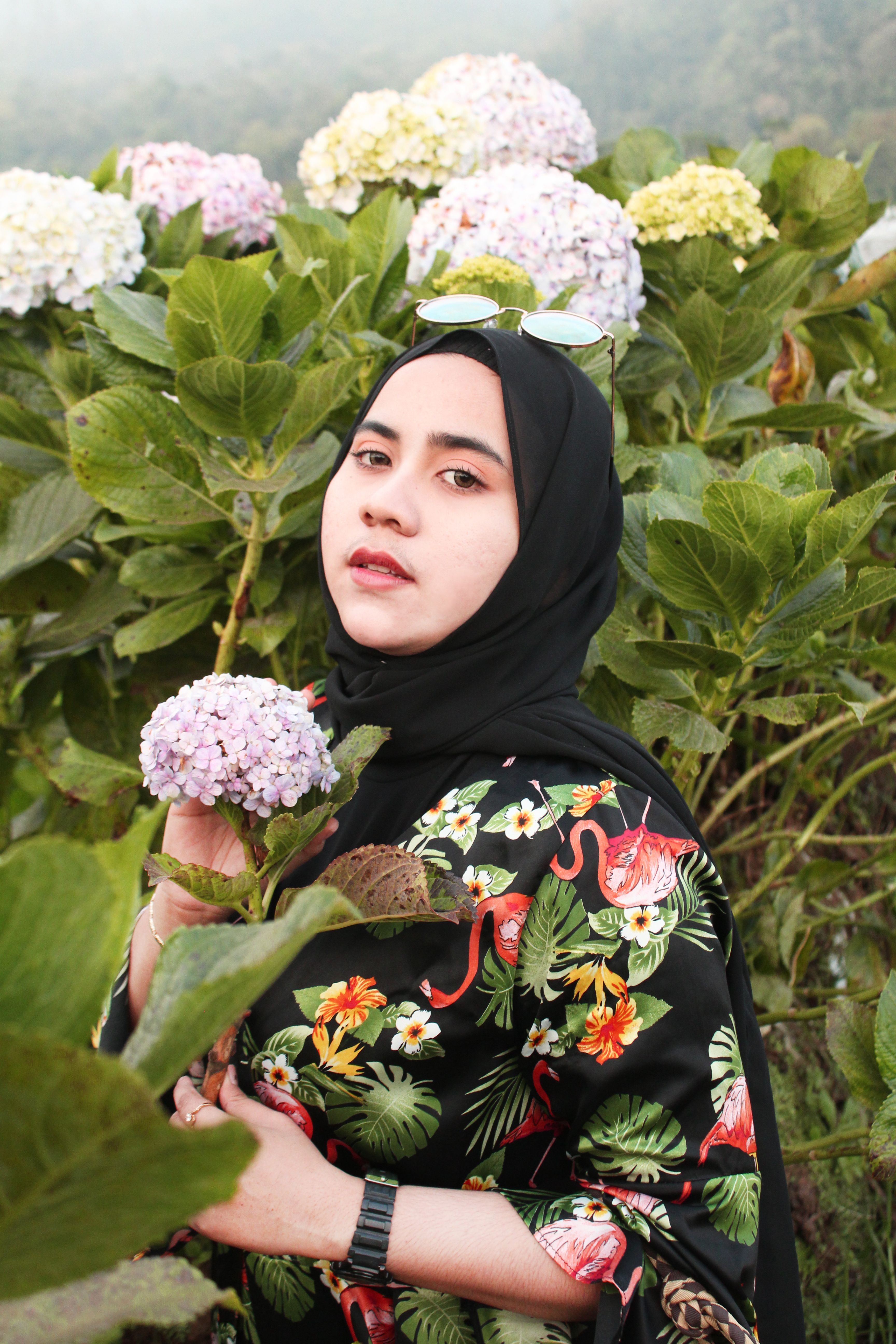 Woman in headscarf posing in a vibrant floral garden, holding a hydrangea.