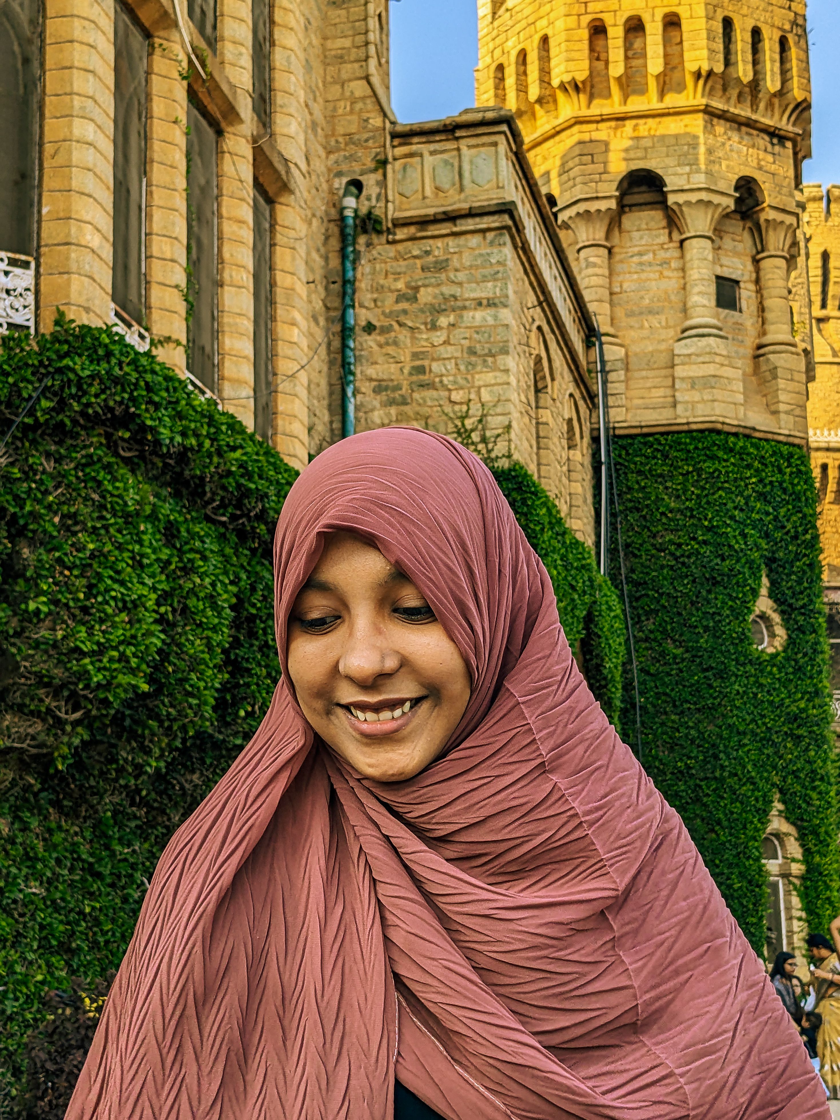 A woman in a hijab smiling warmly while standing near a historic castle.