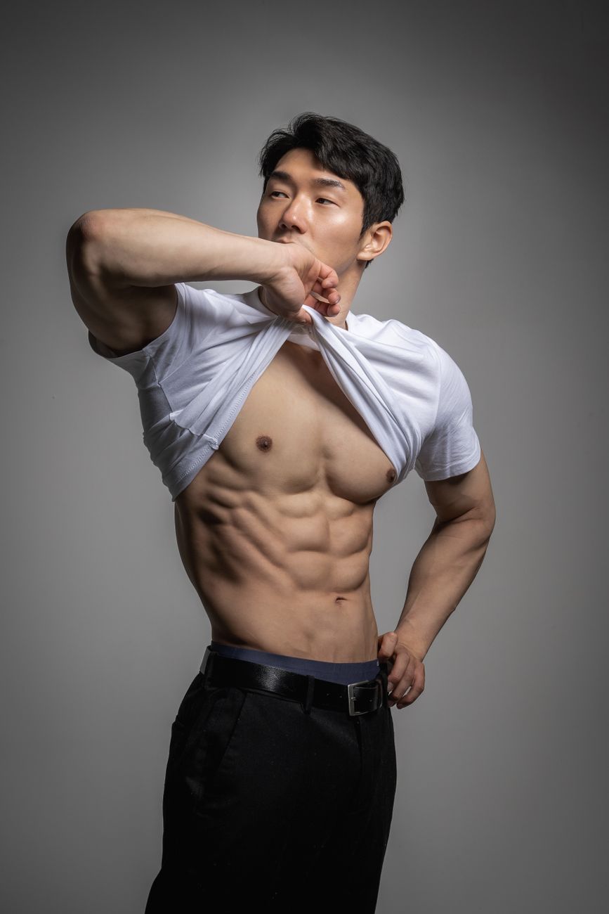 A muscular male model posing in a studio, lifting his shirt to reveal defined abdominal muscles.