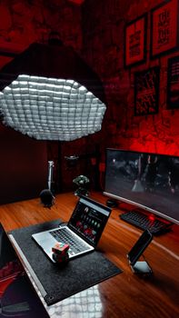 A stylish desk setup with red ambient lighting highlighting a laptop, monitor, microphone, and Rubik's Cube for creative work.