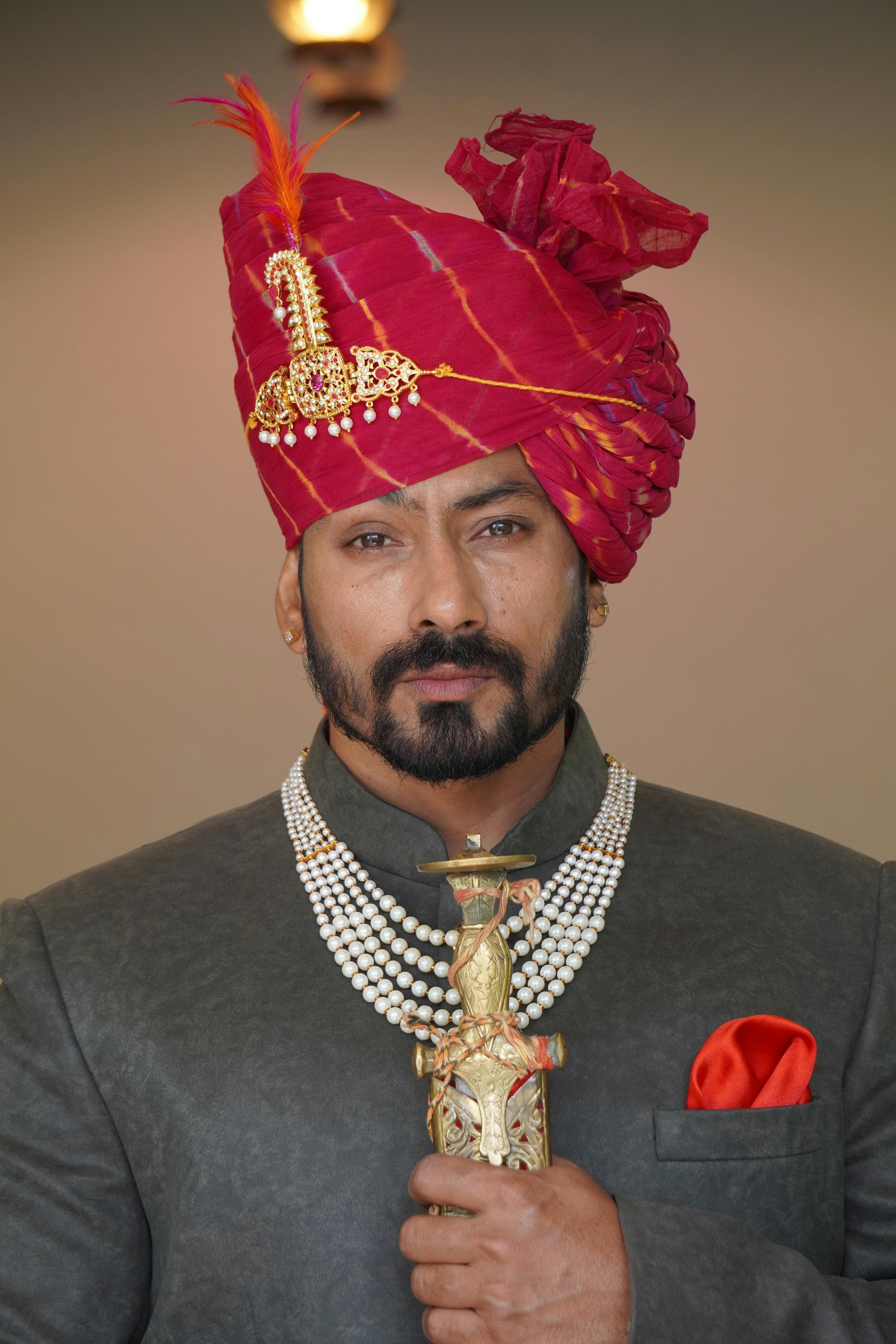 Portrait of a man in elegant traditional attire with red turban and necklace, holding a decorative sword.