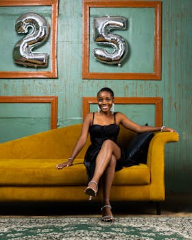 A stylish woman poses on a sofa with '25' balloons on her birthday.