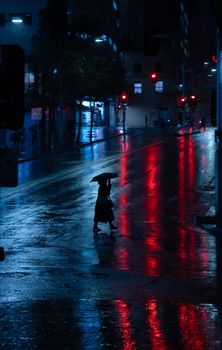 A lone figure with an umbrella crosses a wet, reflective city street at night.