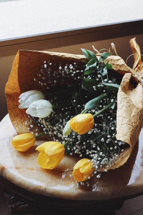 Free Artistic bouquet of yellow and white tulips wrapped in brown paper on a marble table. Stock Photo