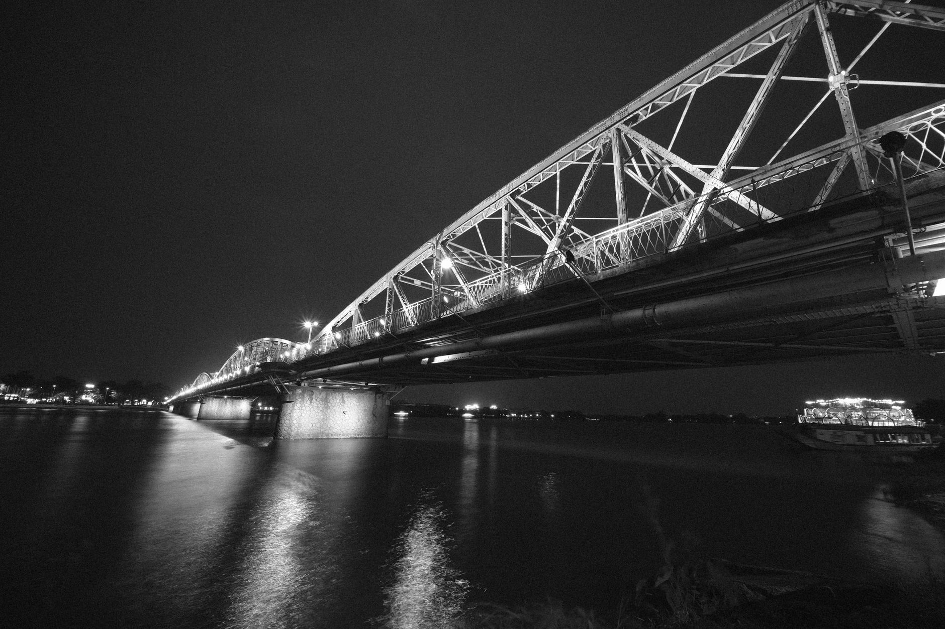 Black and white image of a lit bridge over a calm river at night, Vietnam