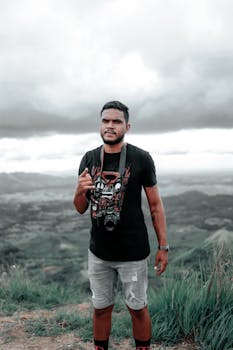 Man holding a camera stands in a rugged mountainous landscape under overcast skies.