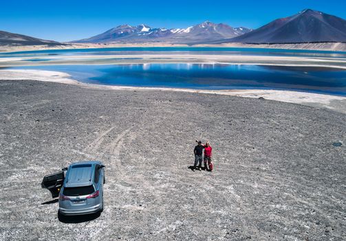 Aerial view of a couple by a scenic lake and mountains with their car, capturing adventure and nature's beauty.