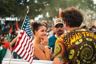 A woman with hands over her mouth experiences emotion during an outdoor patriotic parade with an American flag in view.