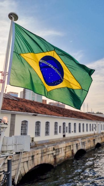 Fotomural Bandera brasileña ondeando cerca de un edificio histórico junto al agua en Río de Janeiro, creando una vibrante escena urbana.