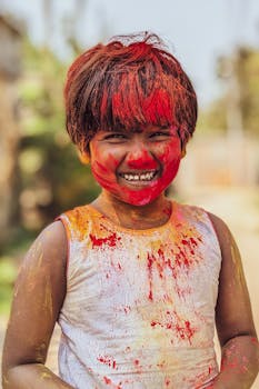 A smiling child with colorful face paint enjoying the Holi festival outdoors.