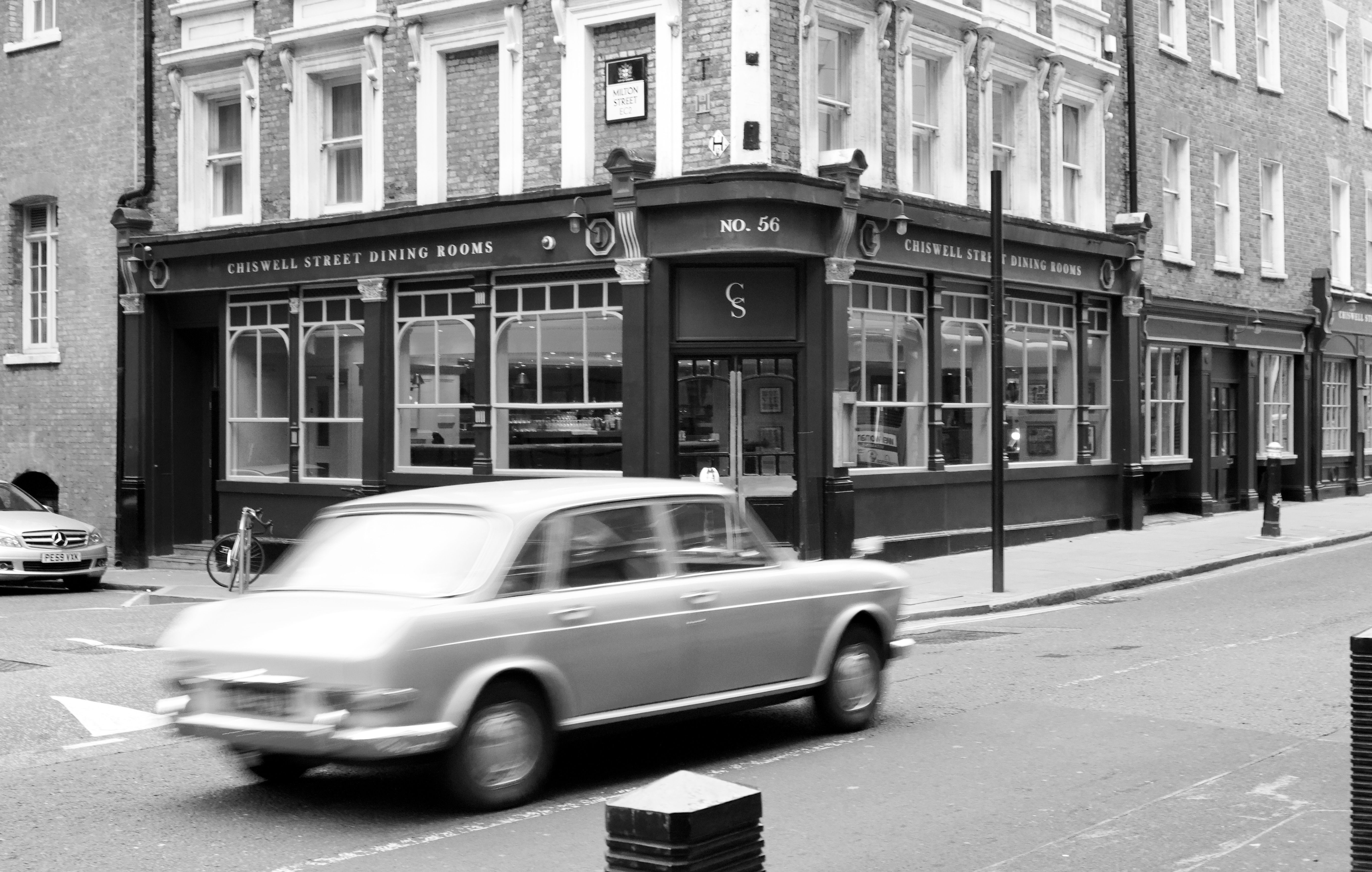 A classic car driving by historic Chiswell Street building in London, captured in black and white.