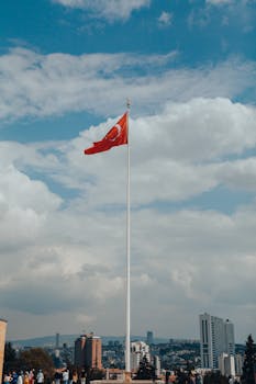 A Turkish flag waves prominently over the urban skyline of Ankara, Türkiye under a cloudy sky.