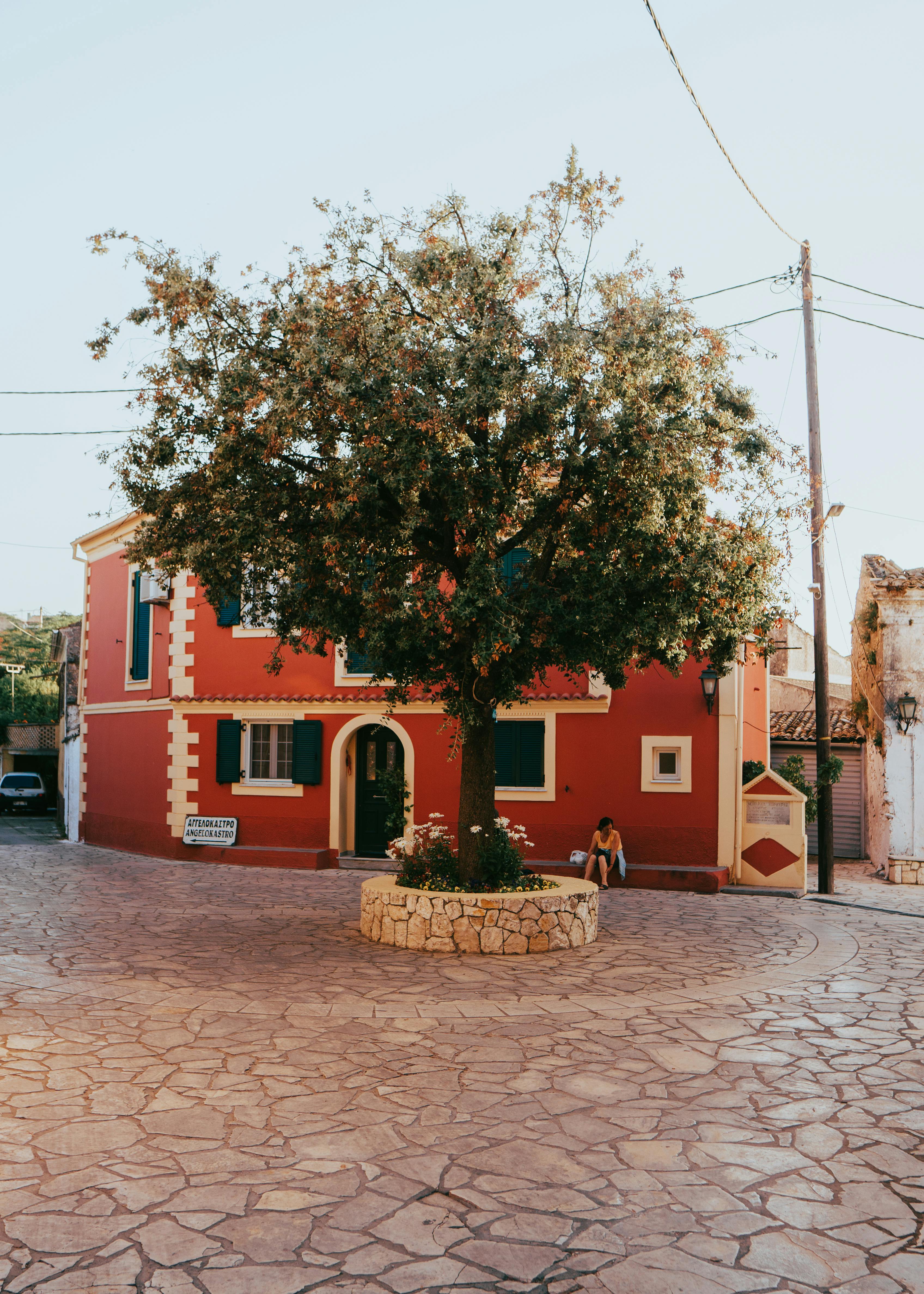 A peaceful square in Kerkyra, Graikija featuring a tree and a vibrant red building under a clear summer sky.