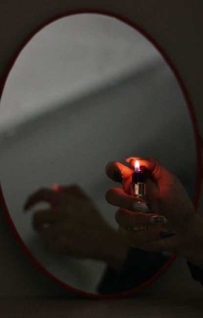 A hand holding a lit lighter is reflected in an oval mirror, creating a dramatic shadowy effect.
