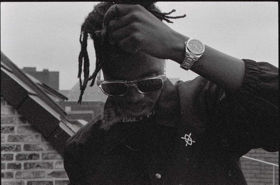Black and white fashion portrait of a man with dreadlocks, sunglasses, and a wristwatch in a cityscape.