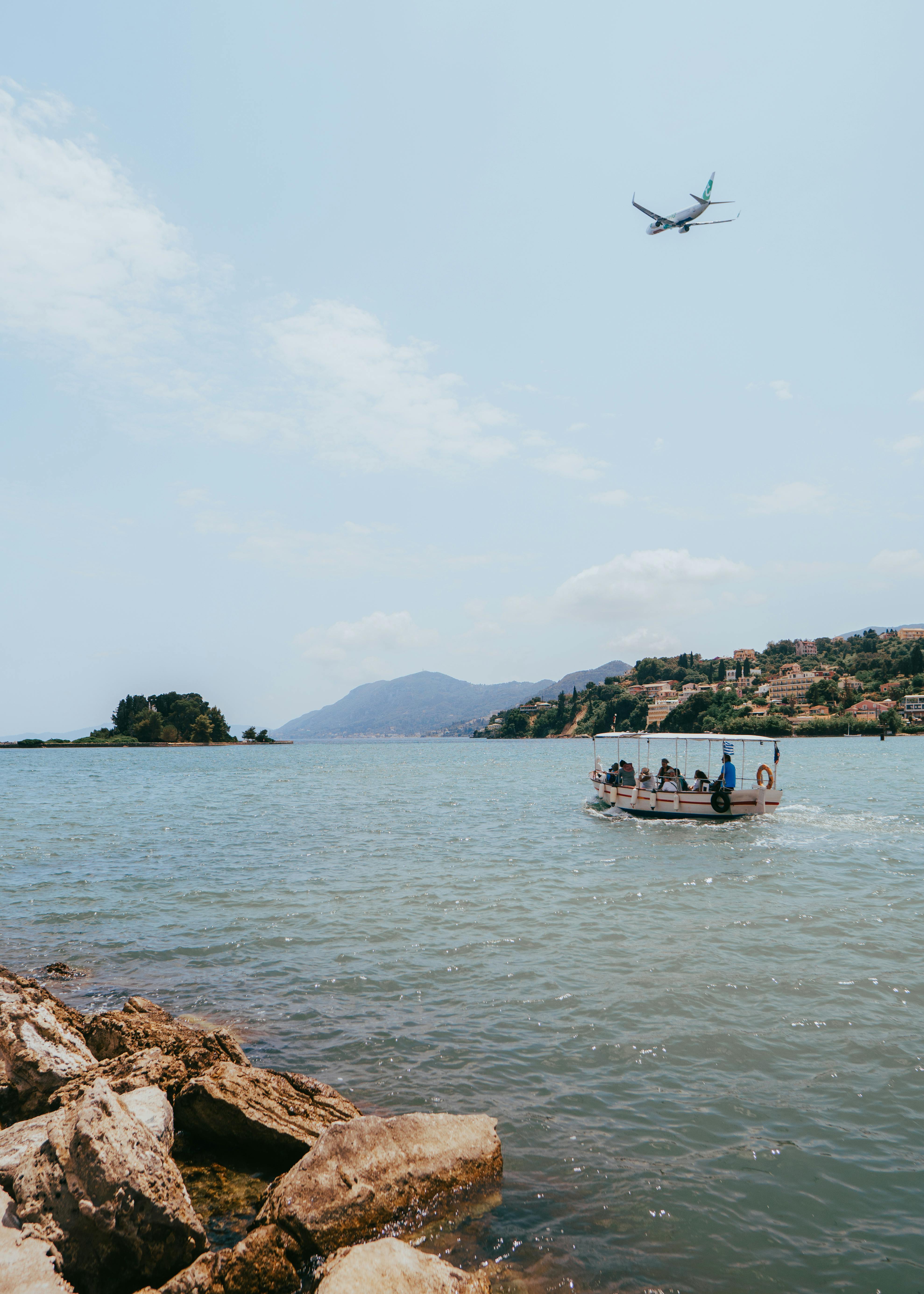 A charming seaside scene in Corfu with a passenger boat and an airplane flying above on a sunny day.