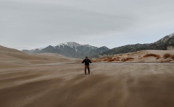 A man stands amidst vast desert dunes with mountains in the background.