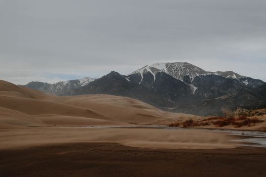 Expansive sand dunes stretch towards snowy mountains under a cloudy sky.