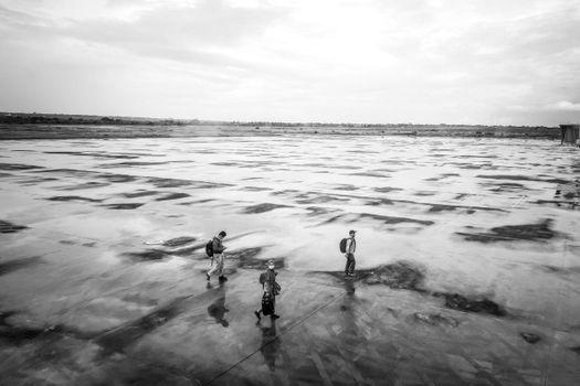 Black and white photo of three people walking across a wet, empty airport runway.