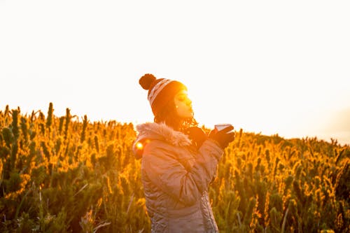 Woman in a Jacket and Hat Standing on a Field at Sunset 