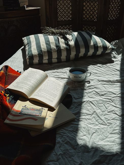 A sunlit bed with striped bedding, books, and a coffee cup creates a serene reading space.