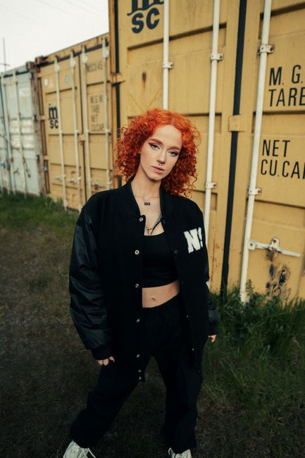 Fashionable redhead with curly hair posing in front of yellow shipping containers.