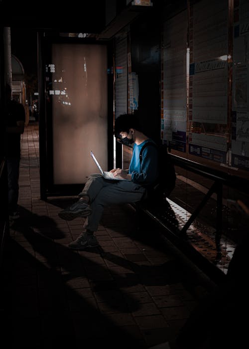 Man in Mask Sitting with Laptop on Bench at Night