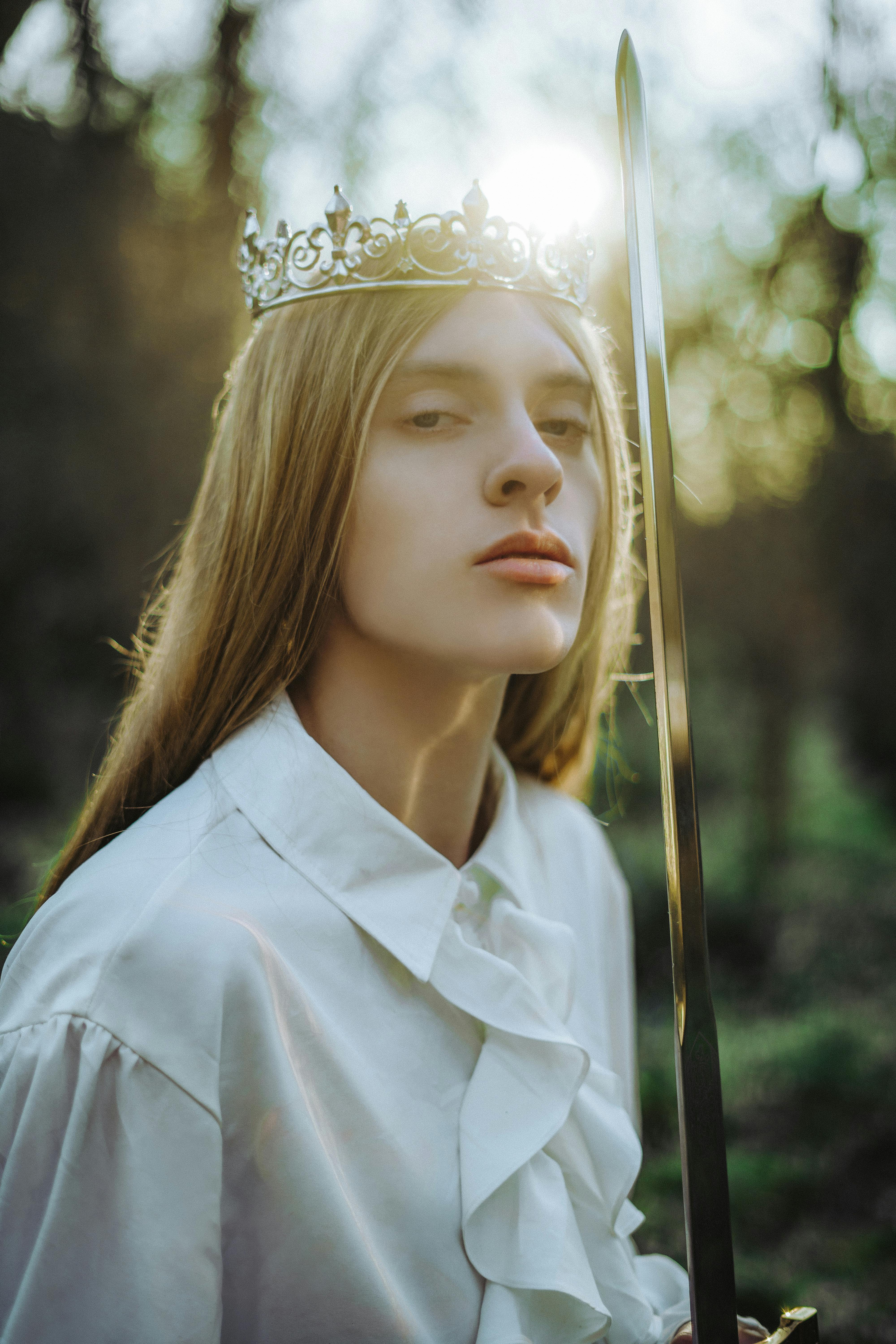 A woman wearing a crown and holding a sword confidently stands in a sunlit forest.