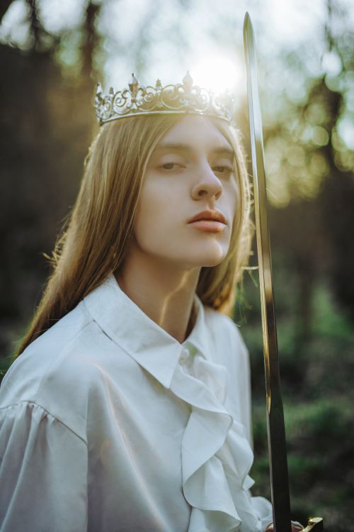 Free A woman wearing a crown and holding a sword confidently stands in a sunlit forest. Stock Photo
