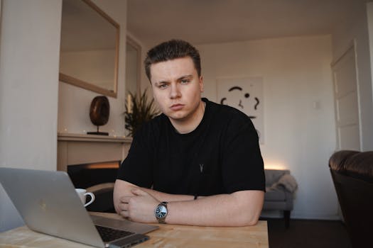 A man working remotely at home, using a laptop at a desk in a cozy living room setting.