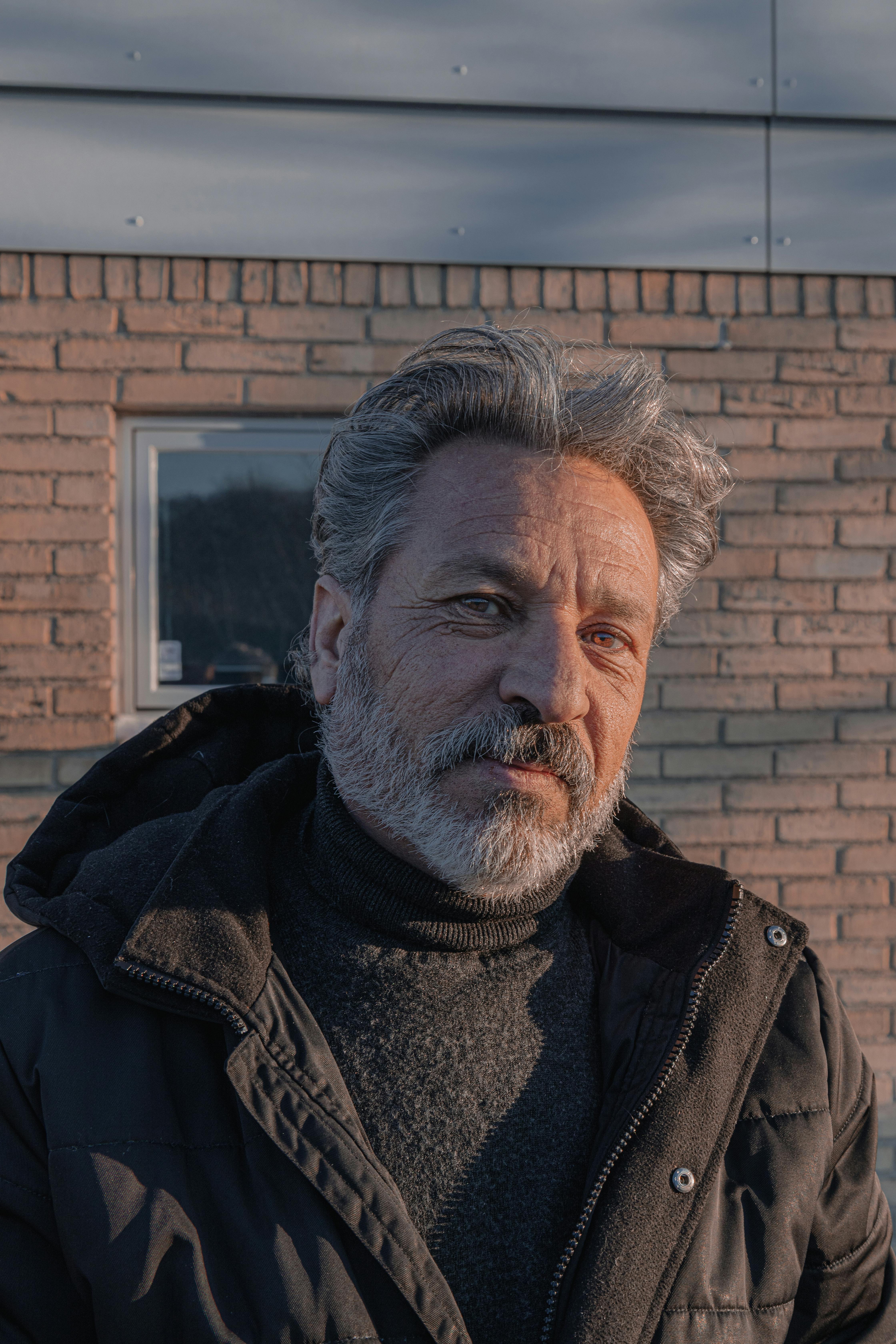 Close-up portrait of a mature man with gray hair and beard, wearing a jacket against a brick wall.