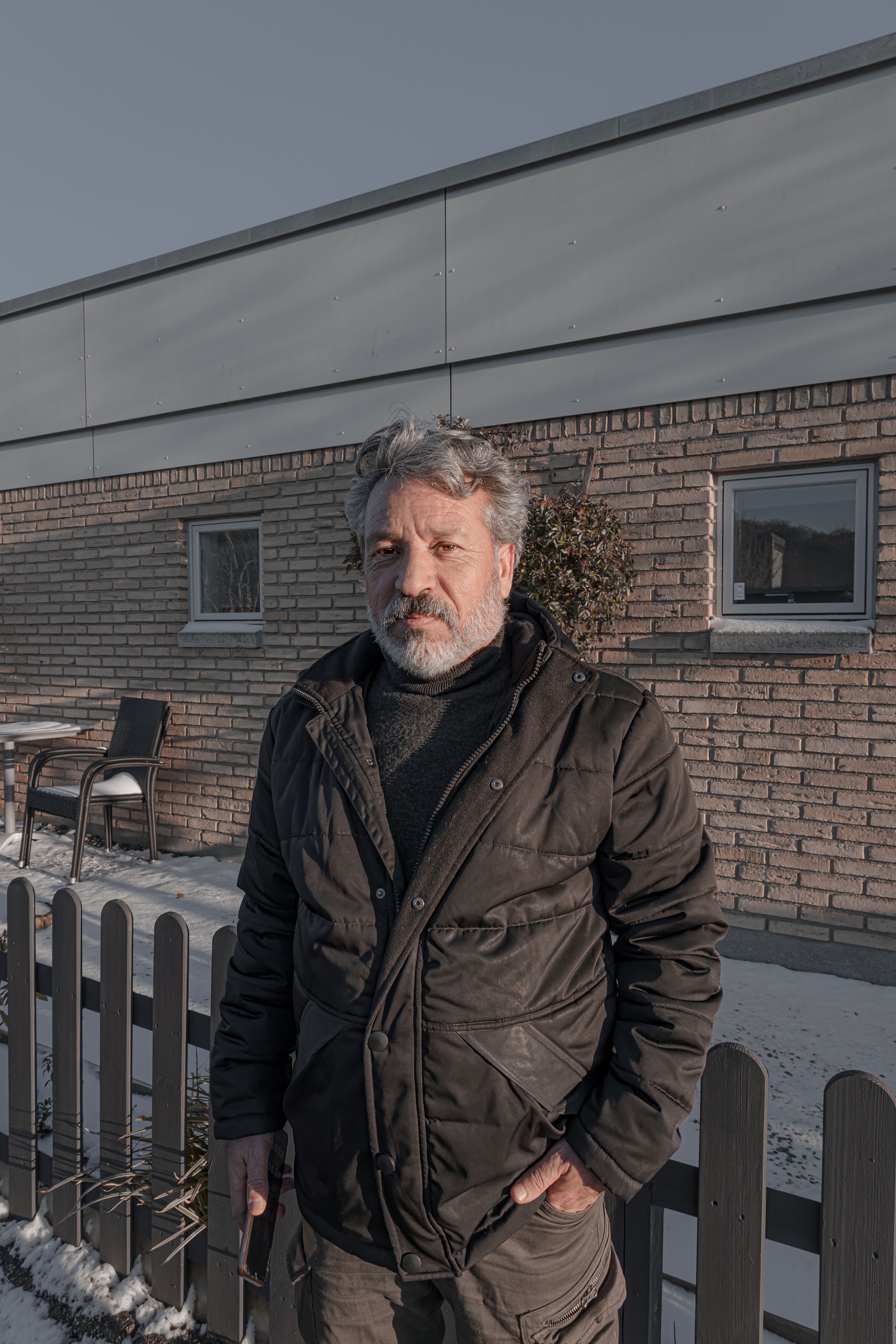 Portrait of an elderly man wearing a jacket, standing by a fence in a winter setting.