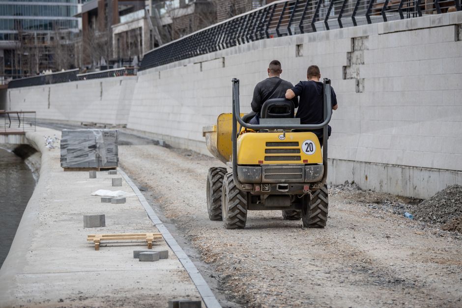 Two construction workers ride a yellow dump truck on a dirt road by a river in an urban area.