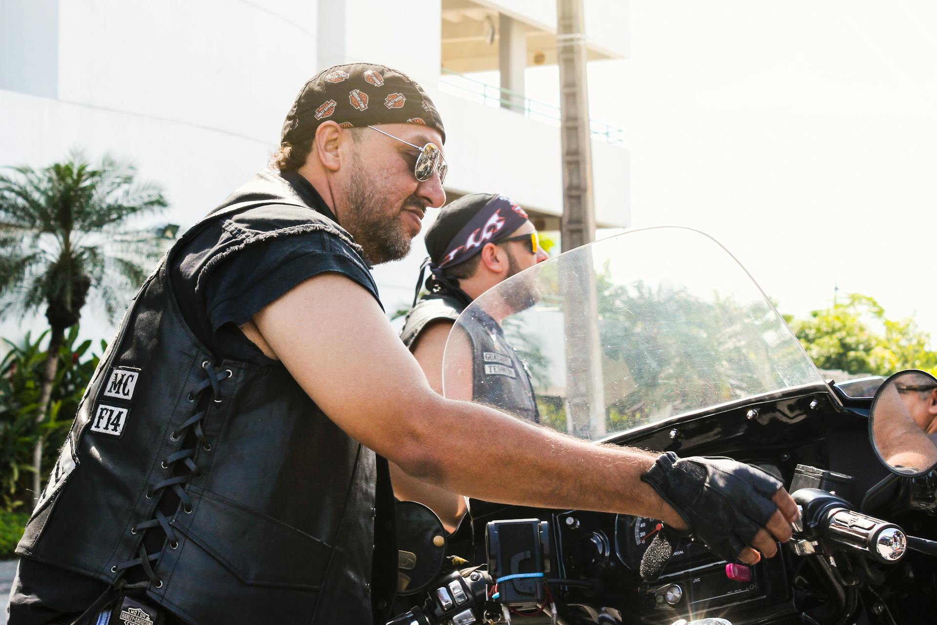 Two male bikers in leather vests riding motorcycles under a bright sun.