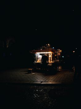 A lone vendor stands at an illuminated food stand on a dark night street, creating a moody ambiance.