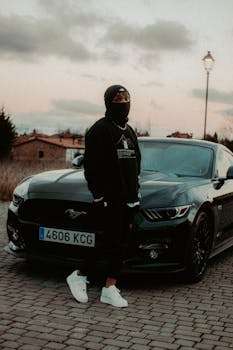 Man in black clothing standing next to a Mustang sports car under a streetlight at dusk.