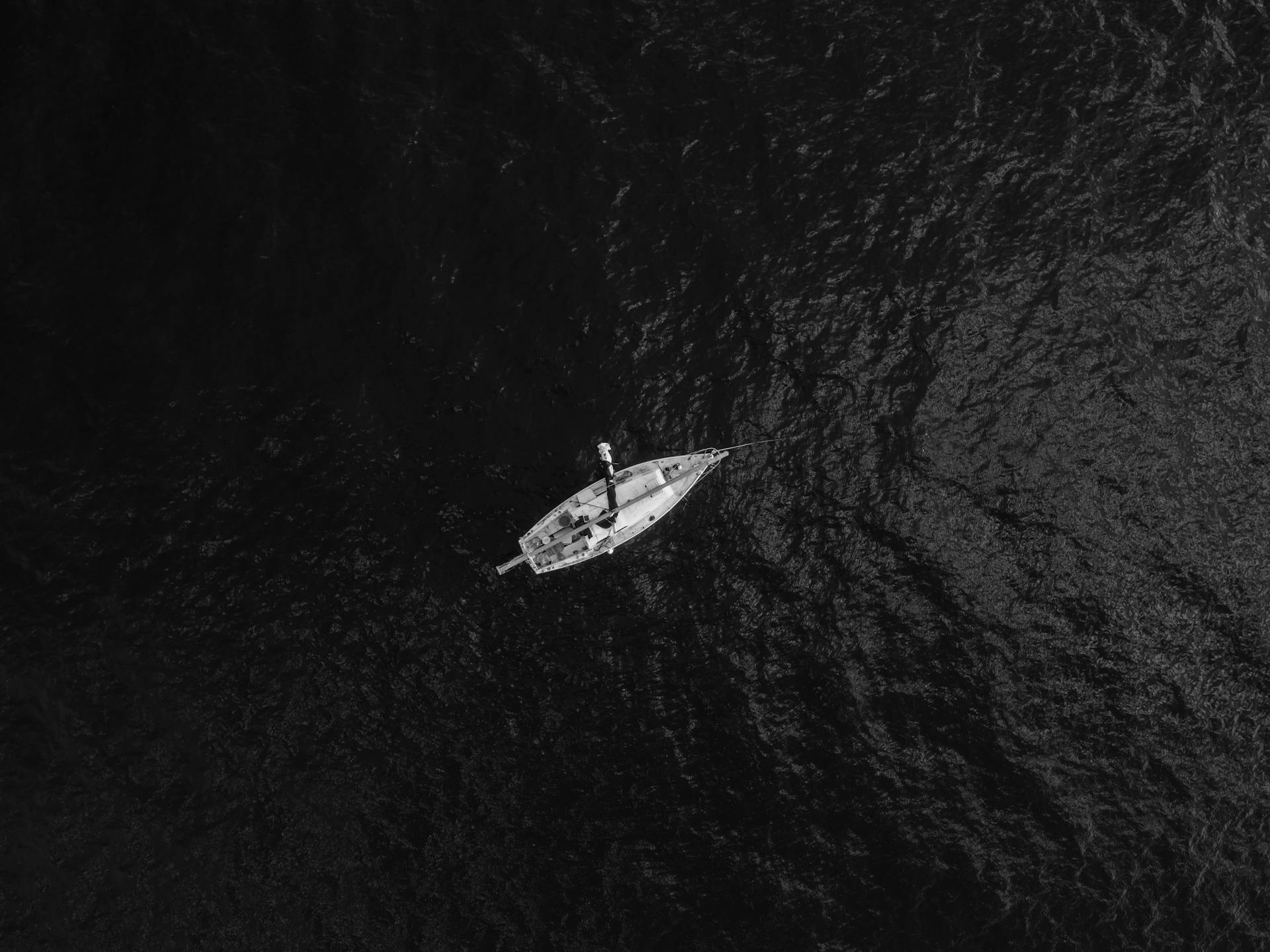 An aerial black and white photo of a lone sailboat navigating vast ocean waters, captured from above.