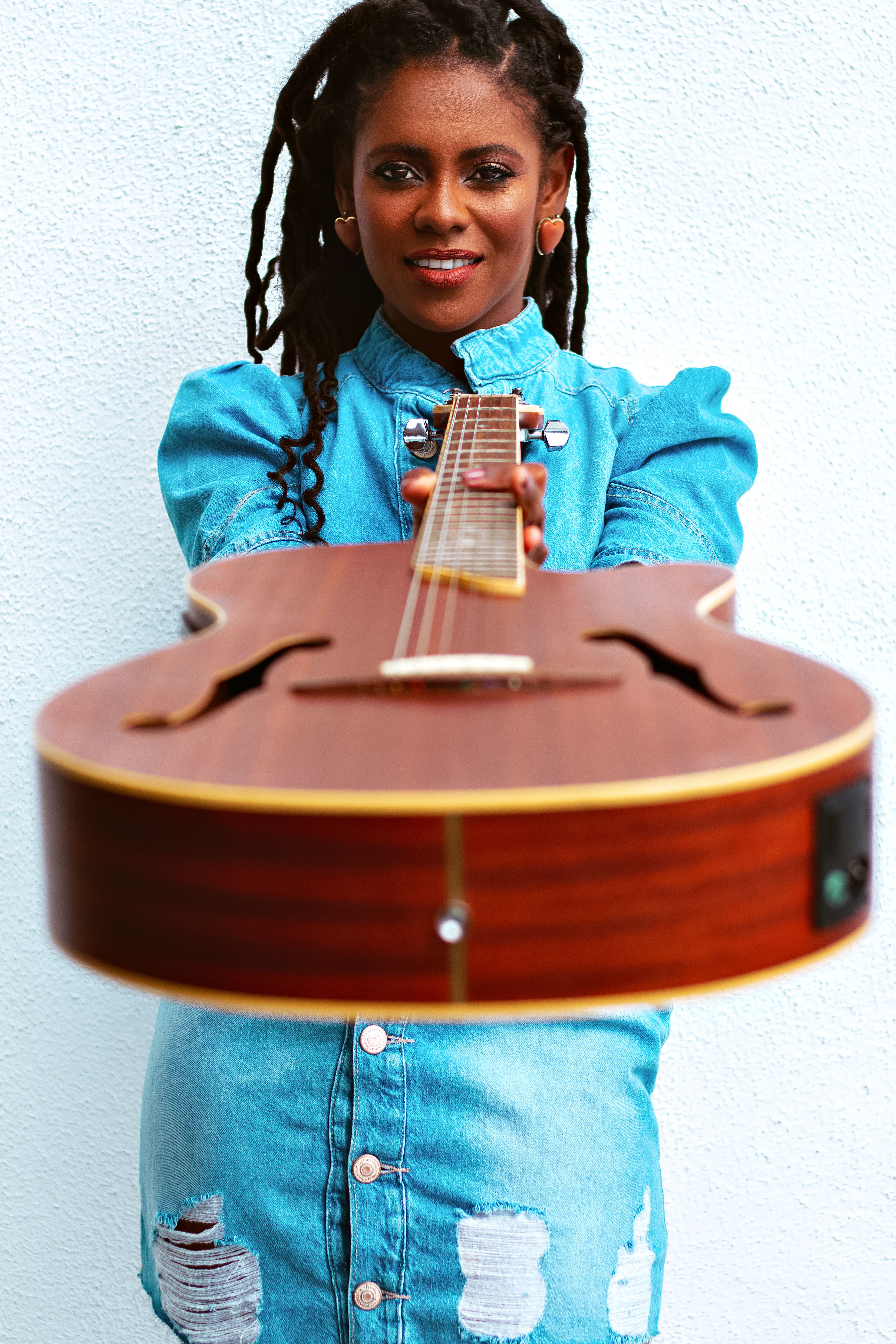 A young woman in denim extends an acoustic guitar forward, smiling warmly.