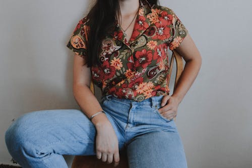 Woman Wearing Multicolored Floral Button-up Shirt and Blue Denim Jeans While Leaning on Wall