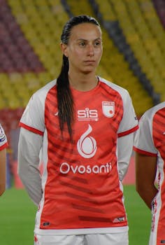 Female soccer player in red and white uniform standing on a football field before a match.