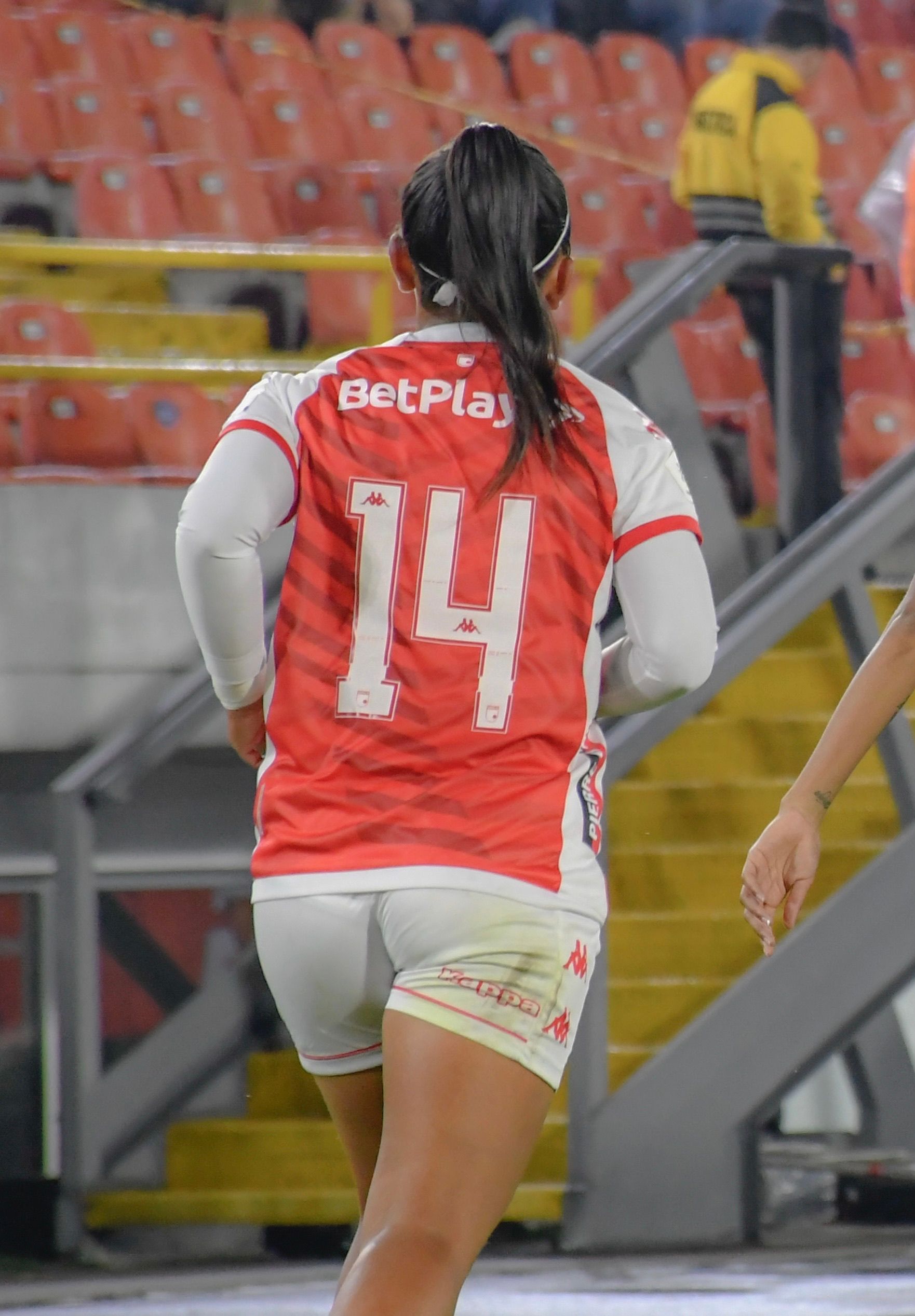 Back view of a female soccer player in action at a stadium, wearing a red jersey.