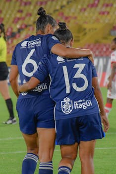 Two female soccer players in blue uniforms embracing on the field after a match.