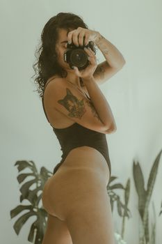 A woman in a black bodysuit captures a selfie in front of a mirror, showcasing tattoos and indoor plants.