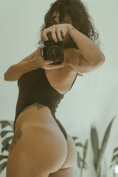 A woman in a black bodysuit takes a selfie using a camera, showcasing tattoos and a mirror reflection.