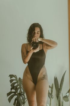 A woman taking a self-portrait in a mirror wearing a black bodysuit surrounded by indoor plants.