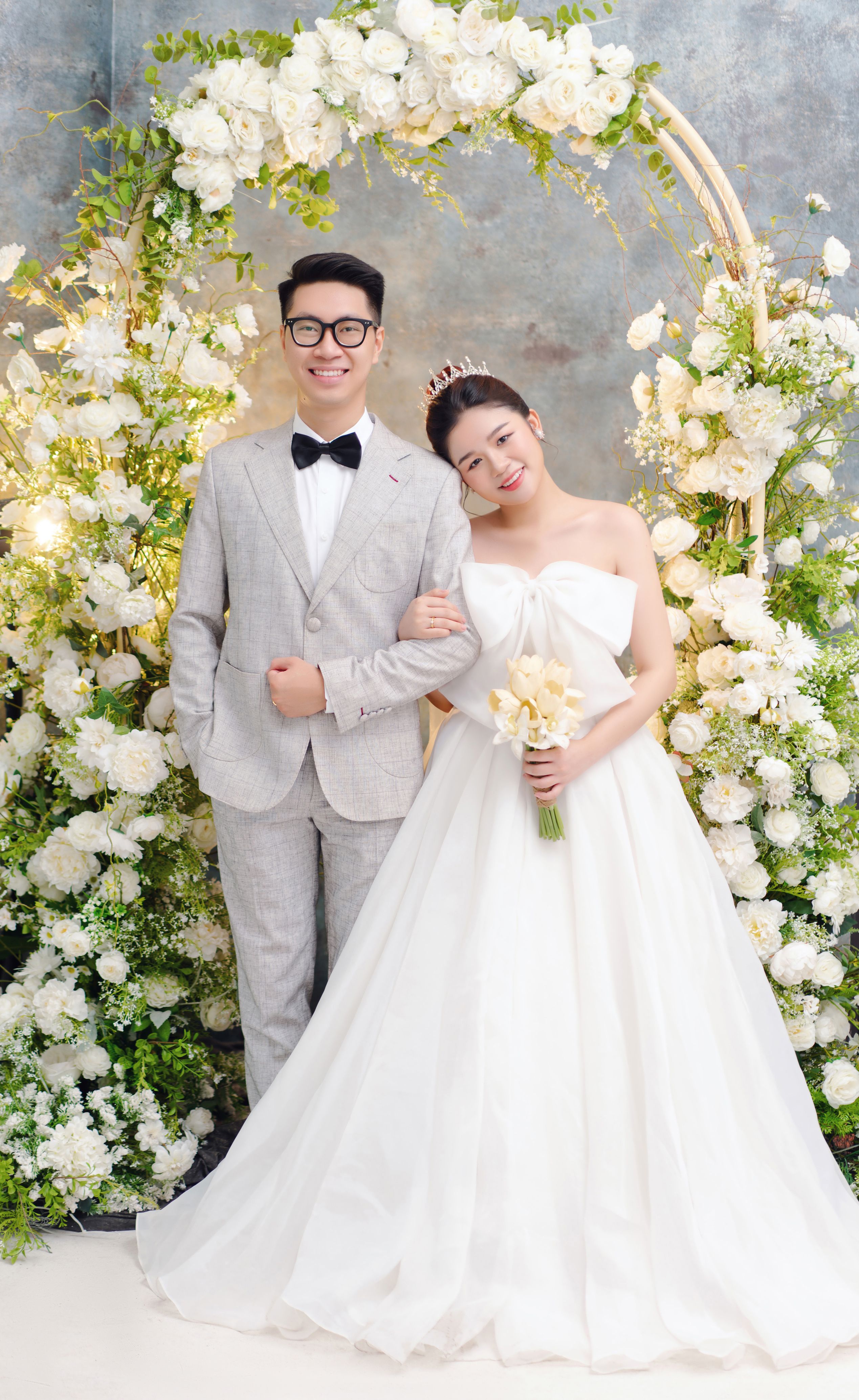 Smiling couple in wedding attire amidst a beautiful floral archway indoors.