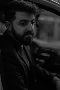 Elegant black and white image featuring a bearded man in a formal suit sitting in a car.
